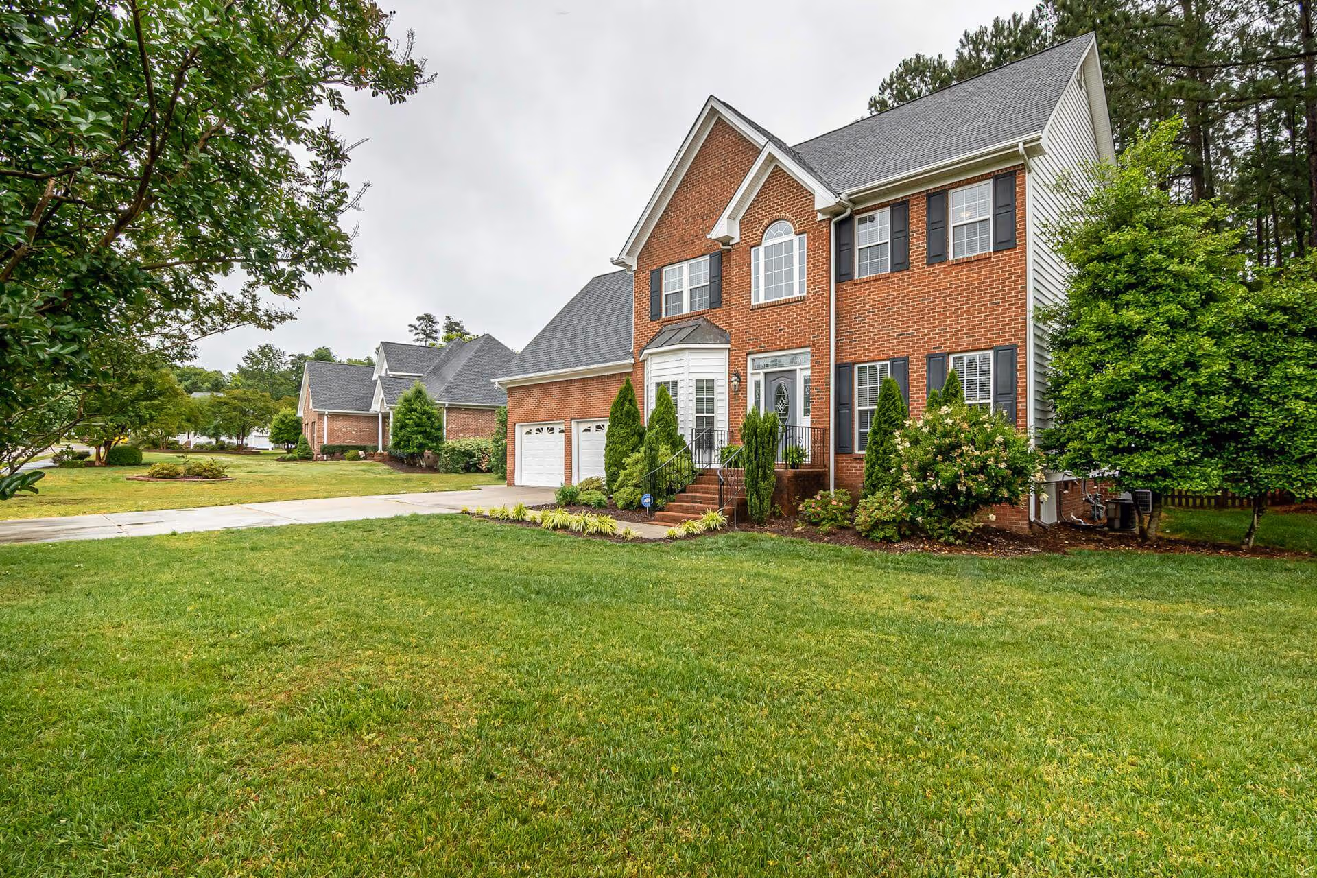 Two-story brick house with large green lawn and surrounding trees on a cloudy day.