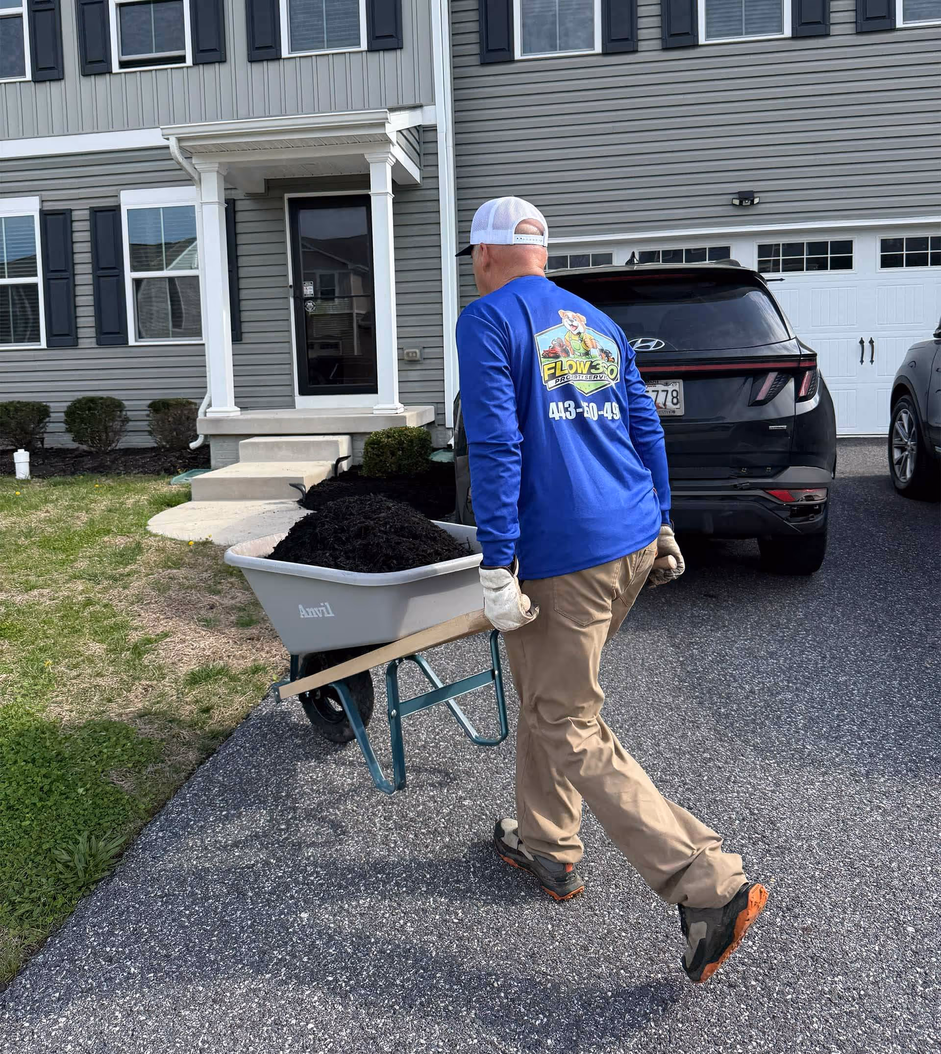Man in blue shirt and khaki pants pushing a wheelbarrow full of black mulch on a driveway near a gray house.