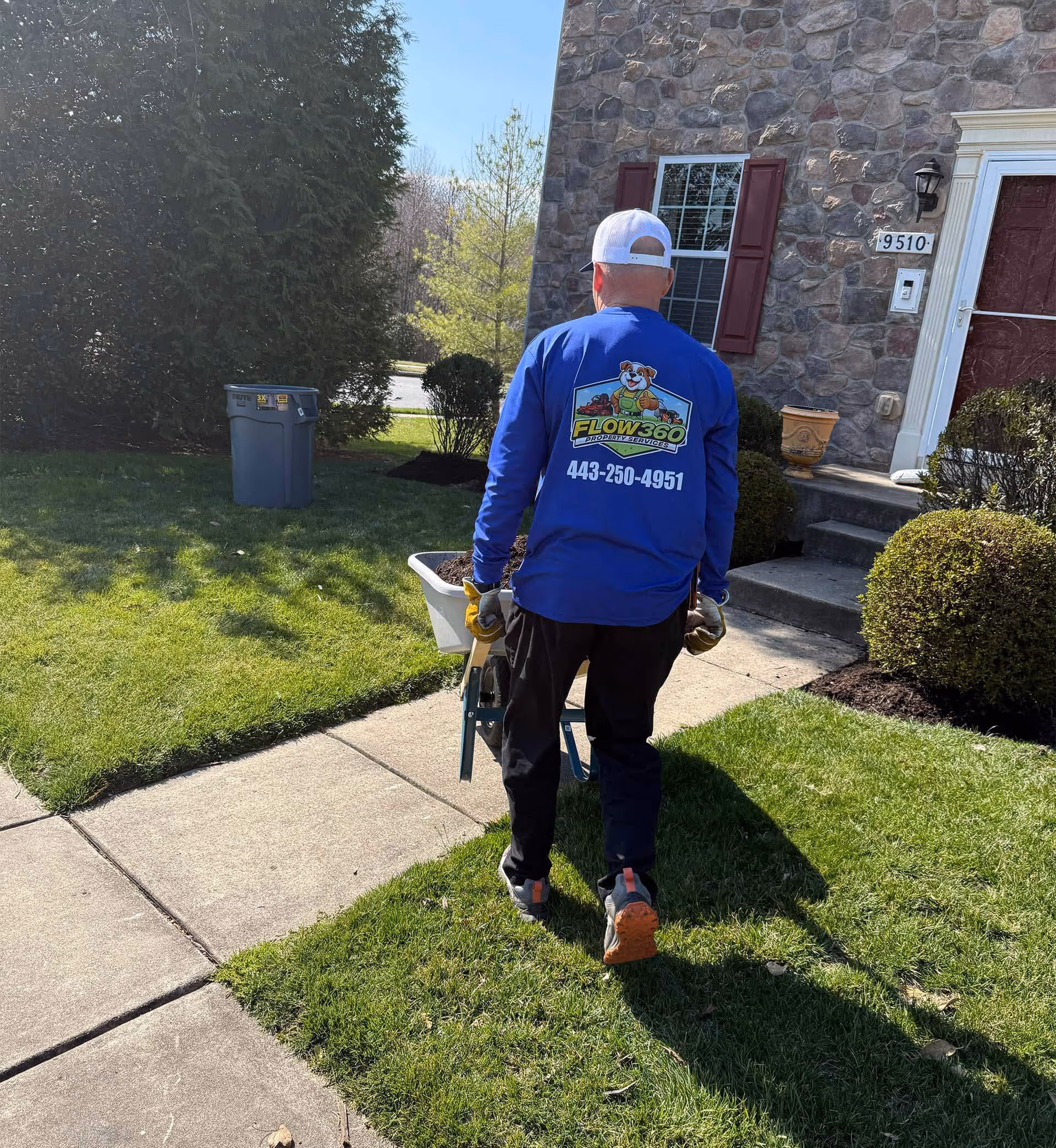 Person wearing a blue Flow 360 Property Services shirt pushing a wheelbarrow with soil on a sidewalk beside a house with stone exterior and red shutters.
