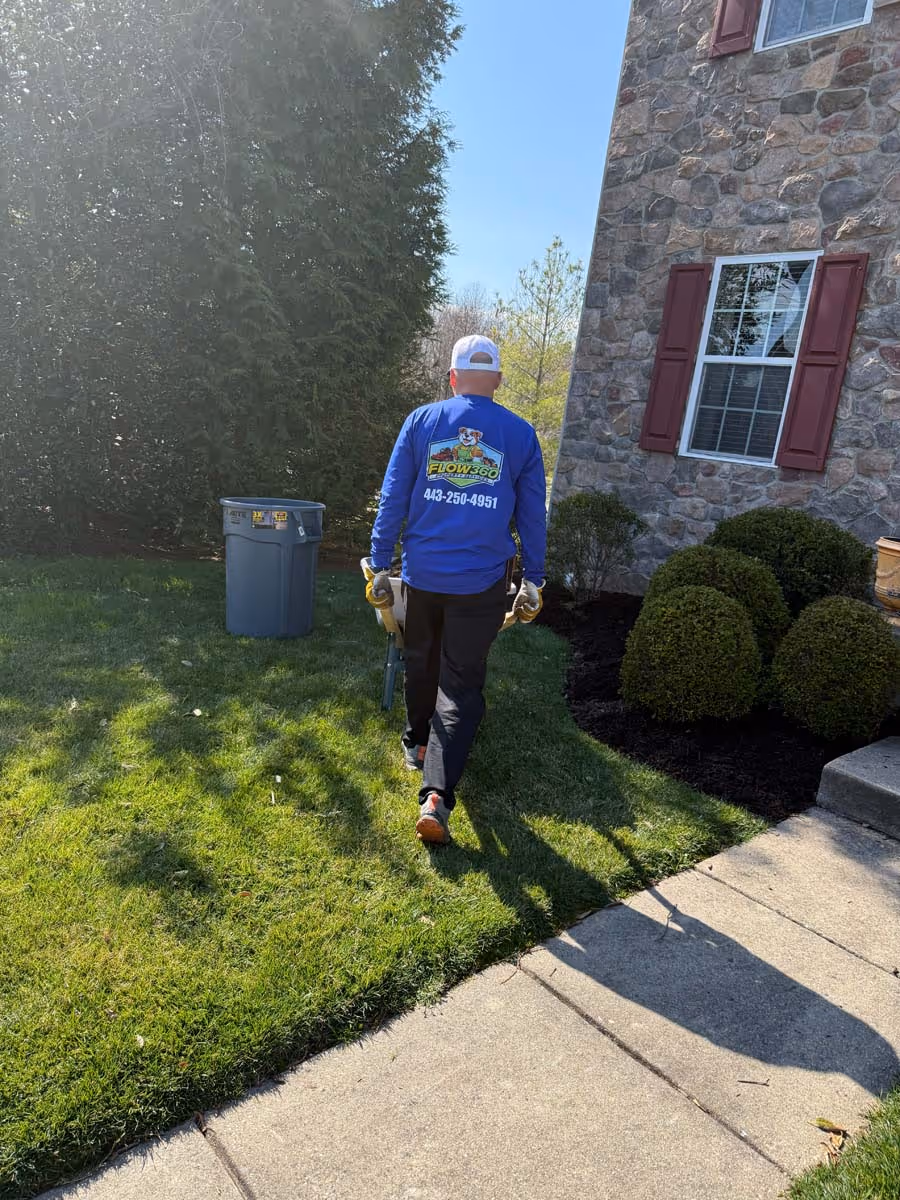 Person in blue shirt with Flow 360 Landscaping logo carrying gardening tools on a lawn next to a stone house and trimmed bushes.