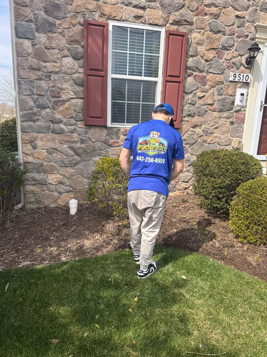 Person in blue FLOW360 shirt and beige pants tending a garden with green grass and bushes in front of a stone house wall with a window and red shutters.