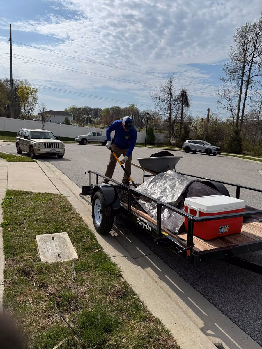Person working with a rake on a utility trailer loaded with soil and a red cooler on a suburban street.